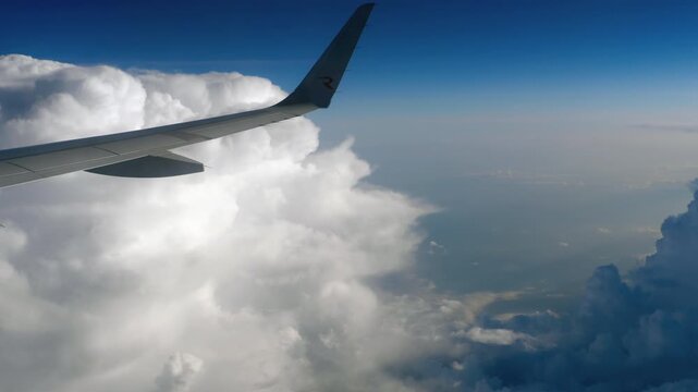 Airplane wing flying above dense white cumulus clouds under a bright blue sky, scenic passenger window view at high altitude