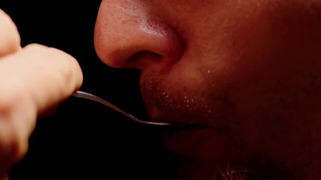 Stubbled Man Tastes Liquid From a Metal Spoon Against Black Background in Extreme Closeup, Showing Food Tasting and Sensory Evaluation