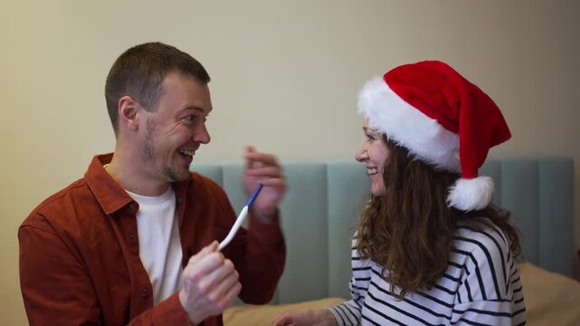 Young caucasian couple celebrating the good news of a positive pregnancy test result, kissing and hugging each other with joy during the christmas holidays while wearing santa hats