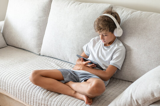 Boy relaxing on a sofa while engaging in online learning, using a digital tablet and headphones for remote education and homeschooling