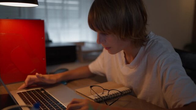 Teen student using a laptop for online learning at home with notebook and eyeglasses on the desk. Digital education, e-learning, remote studying and educational technology concept.