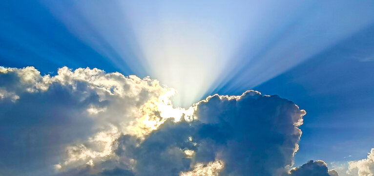 Brilliant sun rays bursting through fluffy cumulus clouds in a clear blue sky, creating a dramatic and serene heavenly atmosphere.