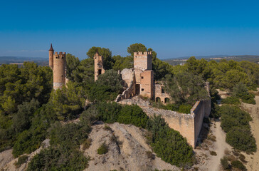 Aerial view of Mas de la Muga, a ruined historicist-style castle near Bellvei, Tarragona, with crenellated brick towers and collapsed walls amid Mediterranean pine forest © IgorsPerspective