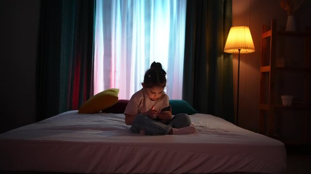 A child sitting on a bed in a dark room in the evening, using a smartphone.