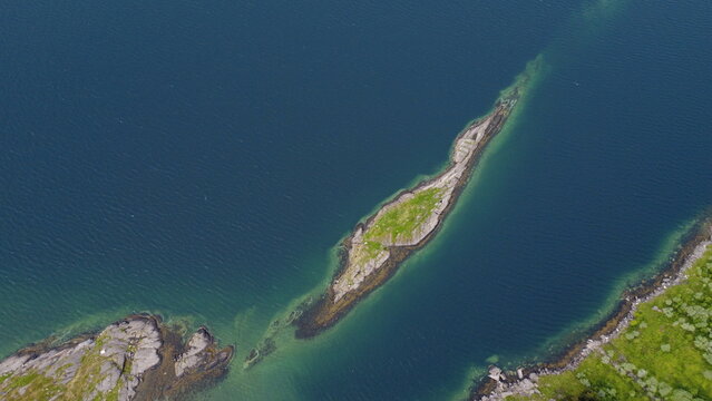 Straight down drone view of a solitary narrow rocky islet with green moss in clear turquoise and deep blue fjord water. Minimalist Scandinavian coastal landscape with water texture.