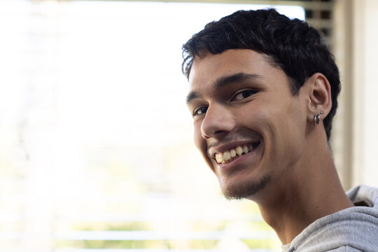 Man smiling, turning to camera at home by blinds, wearing light gray hoodie, earrings, copy space
