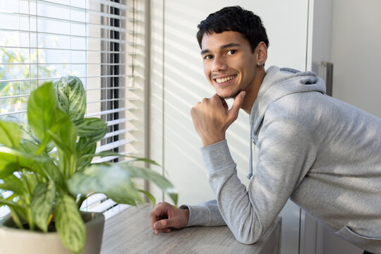 Adult African man leaning on wood table by window with blinds, wearing hoodie, showing hoop earring