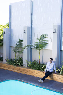 Man sitting on wooden planter bench leaning back, looking right toward turquoise pool, copy space