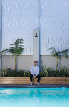 Man sitting on wooden planter bench by swimming pool wearing light gray hoodie and sneakers