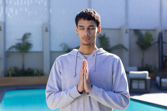 African American man standing pressing palms in prayer by turquoise pool, wearing gray hoodie