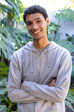 African American man standing crossing arms, wearing hoodie, earrings, tropical foliage in garden
