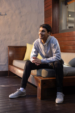 African American man sitting on bench on porch with green pillow wearing gray hoodie, sneakers