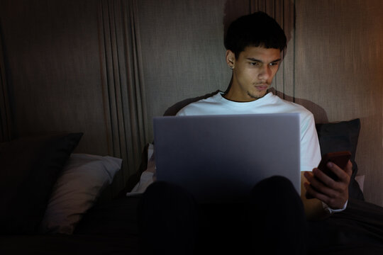 African American man sitting on bed at night holding smartphone while using laptop, white T-shirt