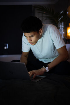 African American man in light tee sitting on bed, leaning forward, working on laptop with lamp