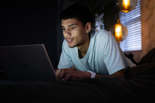 Adult man lying on bed in bedroom, typing on laptop at night with pendant lights, wristband