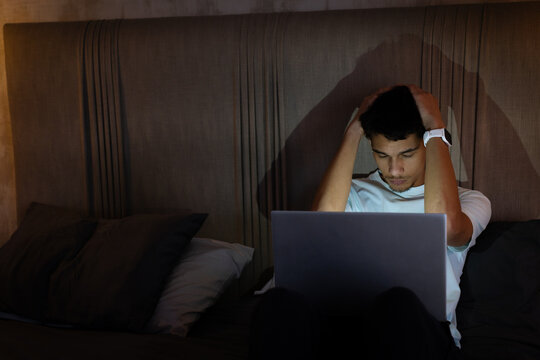 African American man sitting on bed at night, using laptop and rubbing head, copy space