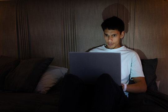 African American man sitting on bed using laptop in dim bedroom with pillows and smartwatch