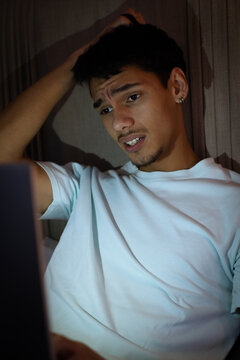 African American man in his twenties rubbing head while staring at laptop glow on sofa