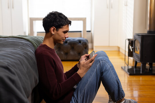 African American man sitting on floor in bedroom leaning against bed holding smartphone