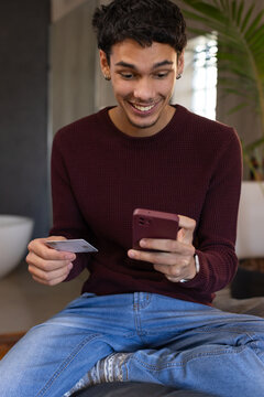 African American man in 20s wearing burgundy sweater holding smartphone and card on cushion at home