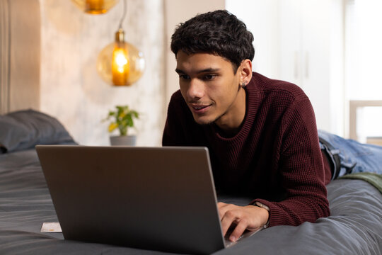 African American man lying on stomach on bed, using laptop, wearing maroon knit sweater and watch