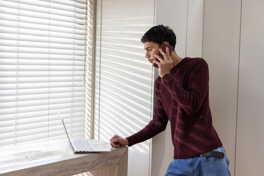 Man in maroon sweater, jeans leaning on ledge by white blinds using smartphone, laptop, copy space