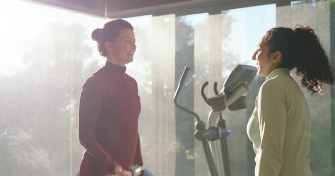 Female friends laughing then curling light dumbbells by windows with elliptical behind for fitness