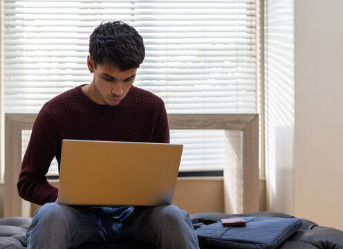 African American man 20s wearing maroon sweater sitting at window using laptop, smartphone nearby