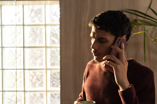 Adult African man standing by window holding phone to ear, green mug, maroon sweater, copy space