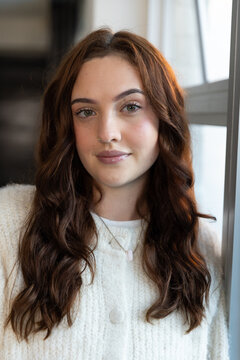 Woman posing near window frame, wearing cream knit sweater, showing pink pendant and nostril hoop