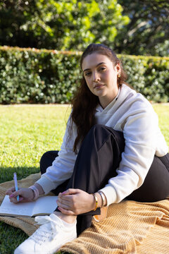 Woman in white hoodie sitting on textured beige blanket on grassy lawn writing notebook holding pen