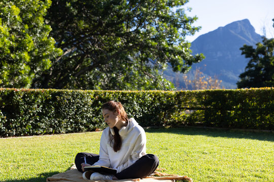 Adult female sitting on beige blanket on lawn, wearing hoodie and headphones, writing in notebook