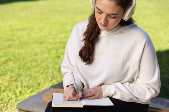 Adult woman in light hoodie writing in notebook with headphones on bench at park, copy space