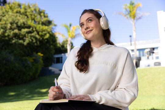 Adult woman sitting on grassy lawn wearing light hoodie and headphones writing in notebook