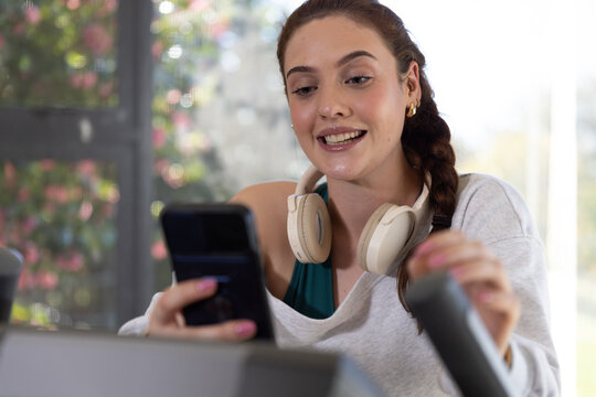 Woman sitting on treadmill holding phone and gripping handle by window, teal sports bra showing