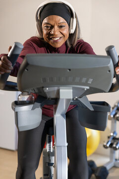 Senior African American woman pedaling on spin bike at home gym wearing maroon top white headphones