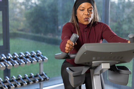 African American woman cycling on bike console in gym, wearing headband and maroon top by weights