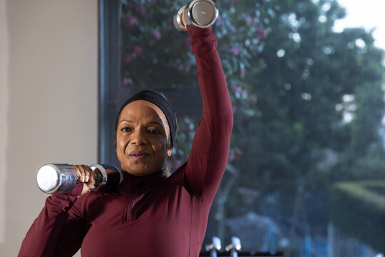 African American woman lifting two chrome dumbbells wearing maroon top and black band in home gym