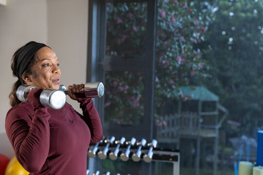 Mature woman holding chrome dumbbells, preparing press in gym by window in maroon top, copy space
