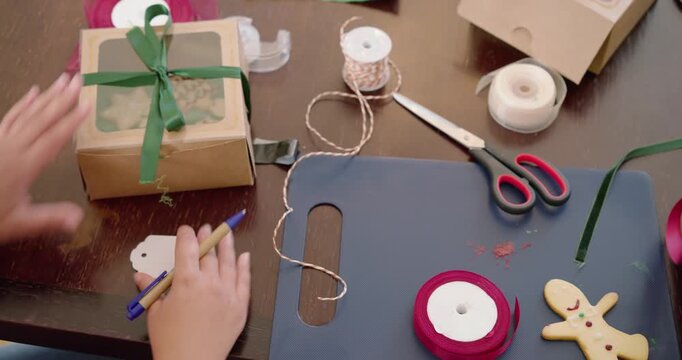 Woman preparing gift at home after seeing bakery box, tying ribbon on kraft box, writing tag
