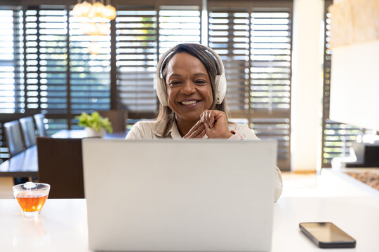 Senior African American woman wearing white headphones, typing on laptop at home with tea