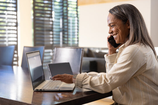 Mature African woman in shirt, watch, holding smartphone, using laptop at home table, copy space