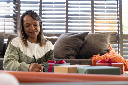 Mature adult African woman writing card among wrapped gifts near gray sofa holding pen, copy space