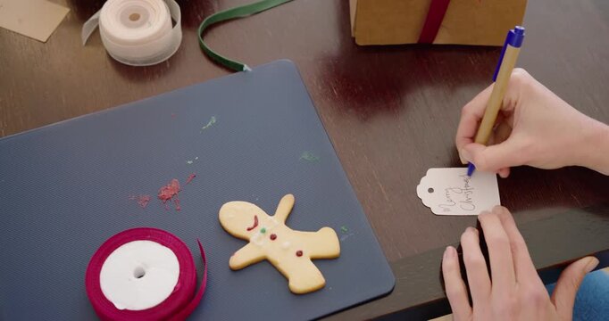 Yellow-sleeved female hands writing scalloped gift tag on dining table, preparing gift with red bow