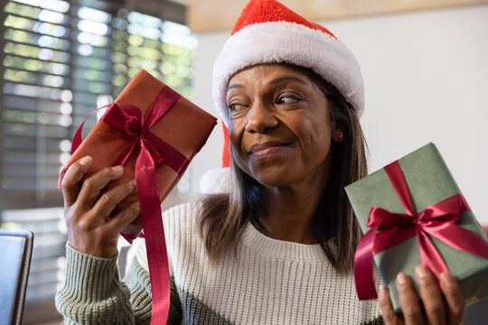 Senior African American woman wearing Santa hat and knit sweater holding two ribboned gifts at home