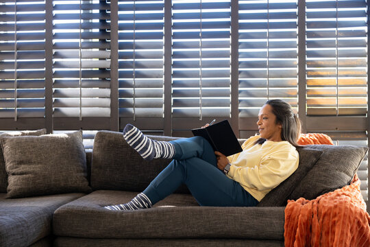 Mature African American woman on gray sectional sofa writing in black notebook with pen, copy space