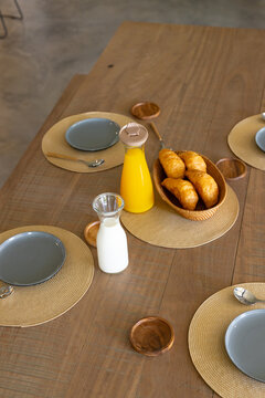 Wooden table is hosting croissant basket, milk carafe and sealed orange juice in dining area