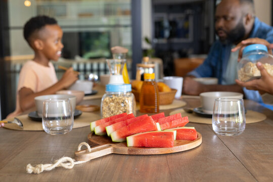 African American father and child sitting at dining table, sharing watermelon from wooden board