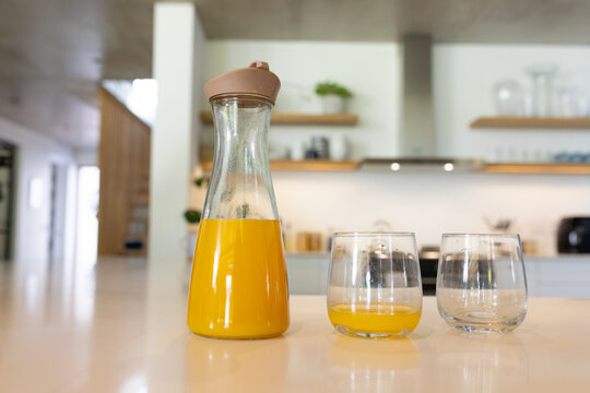 Glass carafe sitting two-thirds full of orange juice on kitchen counter, with two stemless glasses