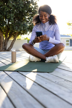 African American woman sitting on green mat on deck, scrolling phone, wearing light purple top